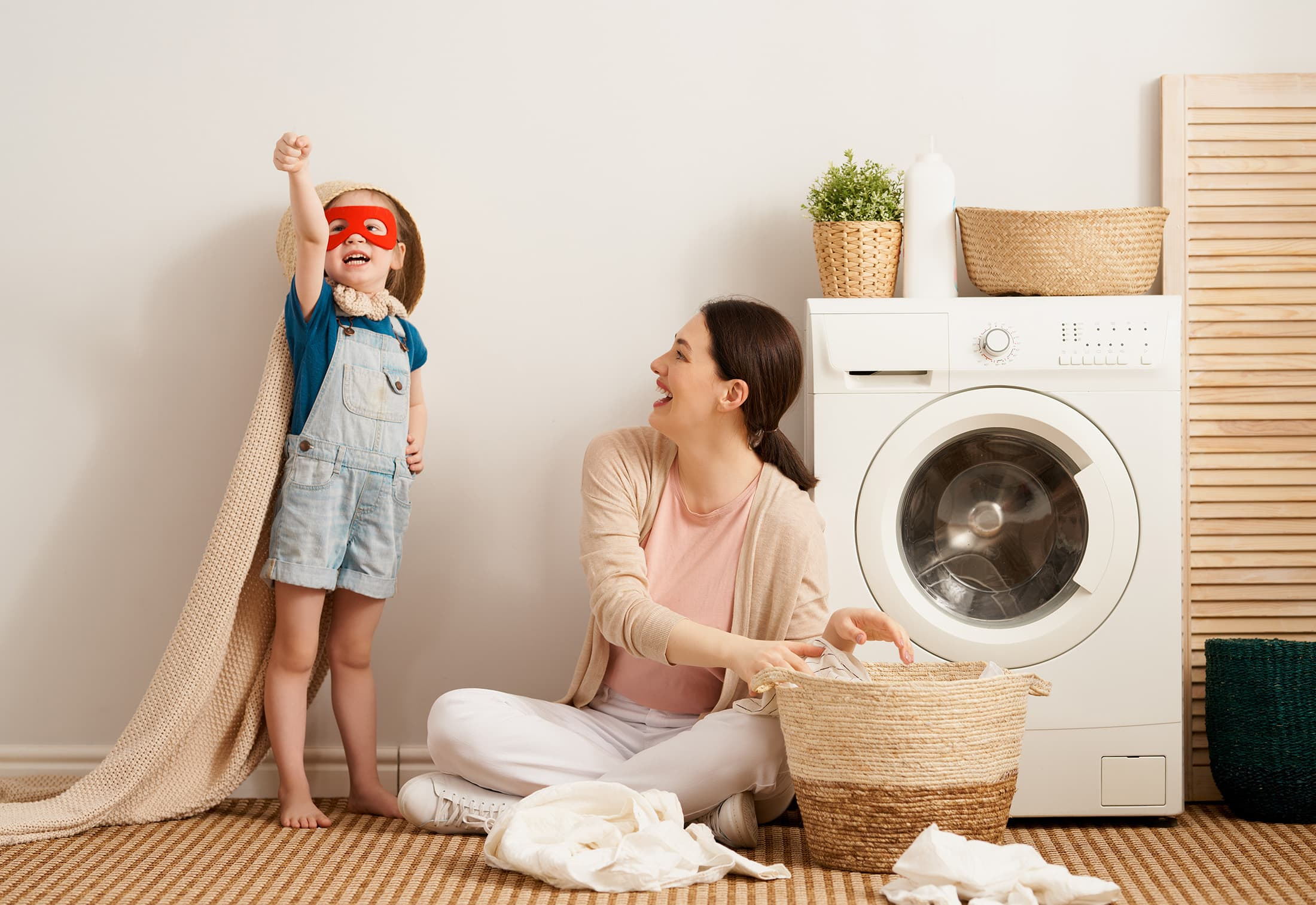 Eine Frau sitzt mit ihrer Tochter vor der Waschmaschine. Das Mädchen spielt Superman mit einer Decke.