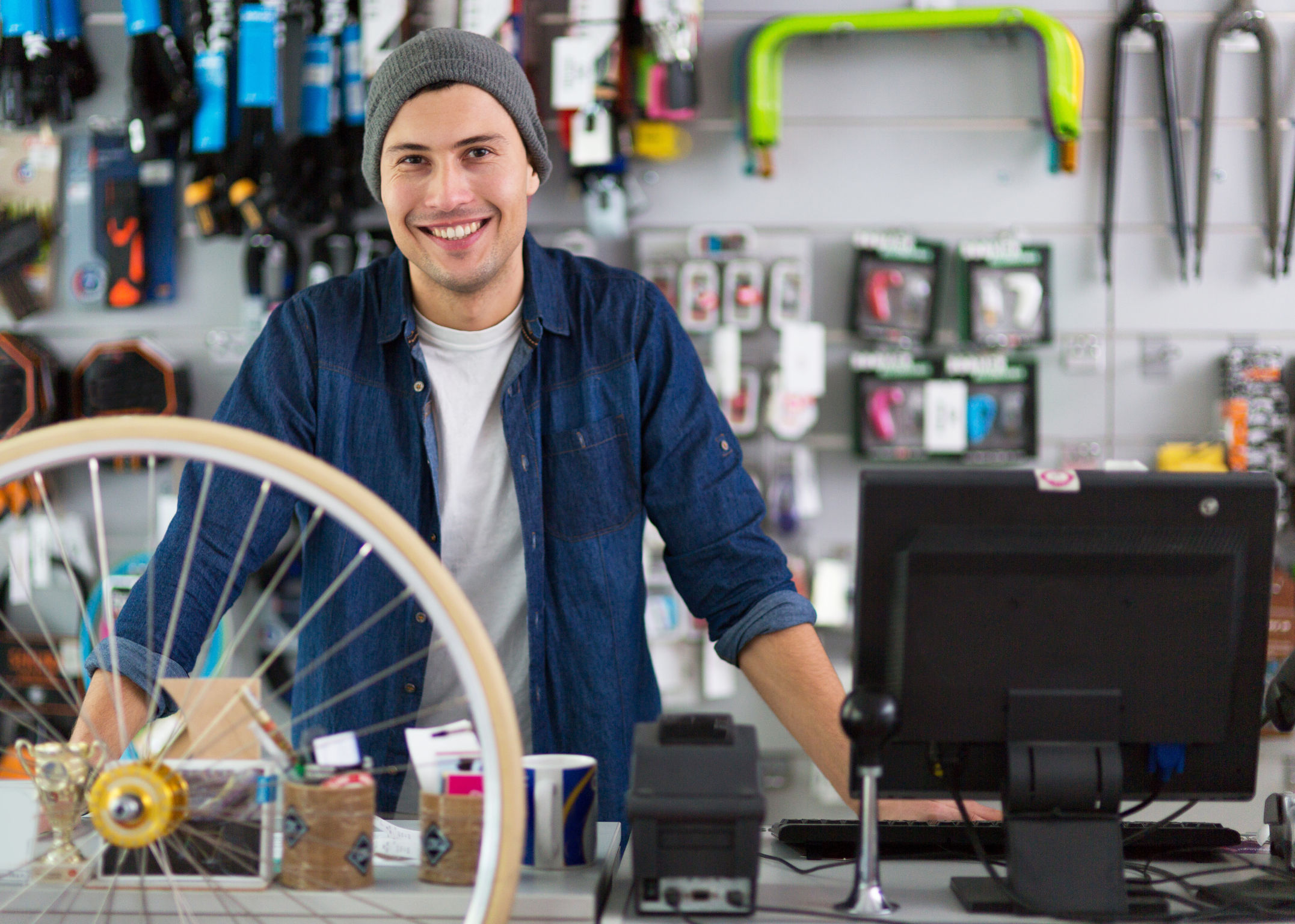 Ein Mann steht in einem Fahrradgeschäft lächelnd hinter dem Tresen. Im Vordergrund ist ein Reifen und ein Desktop zu sehen.