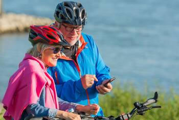 Zwei ältere Menschen am Wasser mit Fahrrädern und Fahrradkleidung schauen auf Handy