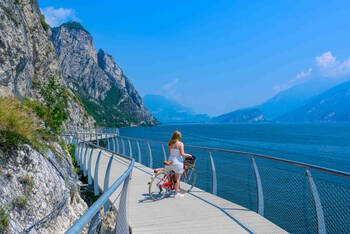 Frau mit Rad steht auf einer Brücke vor See in bergiger Landschaft