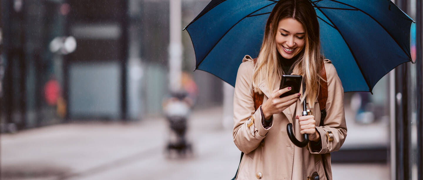 Eine junge Frau im beigen Trenchcoat mit Regenschirm schaut lächelnd auf ihr Smartphone