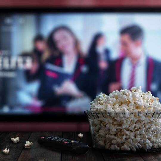 Kosten für Netflix steigen Fernseher mit Netflix im Hintergrund, im Vordergrund sind Popcorn und Fernbedienung
